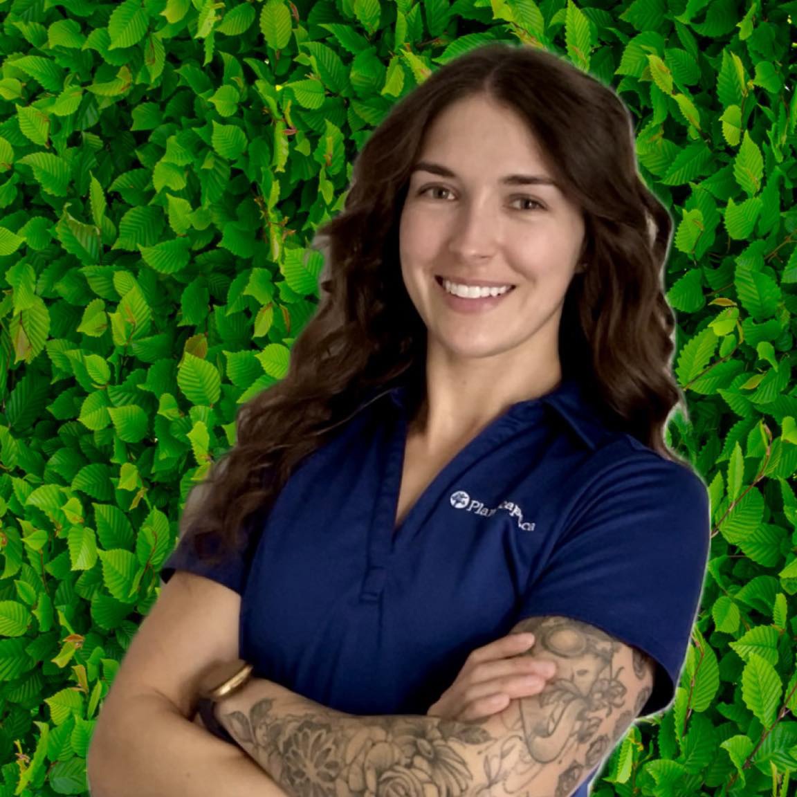 A person with long hair and a tattooed arm smiles, wearing a blue shirt against a green leafy backdrop. They appear friendly and professional.