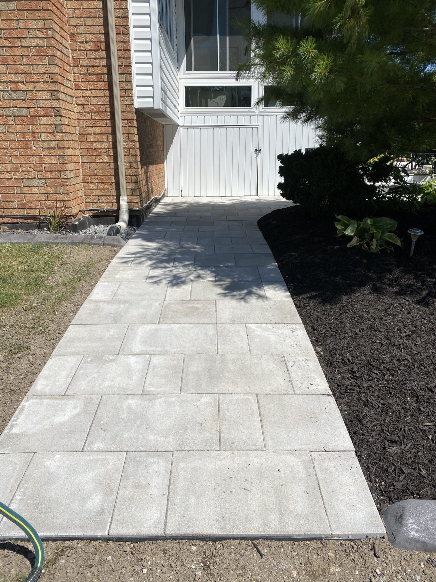 This image shows a neatly paved pathway leading to a side entrance with white siding. A large brick wall and lush greenery border the path.