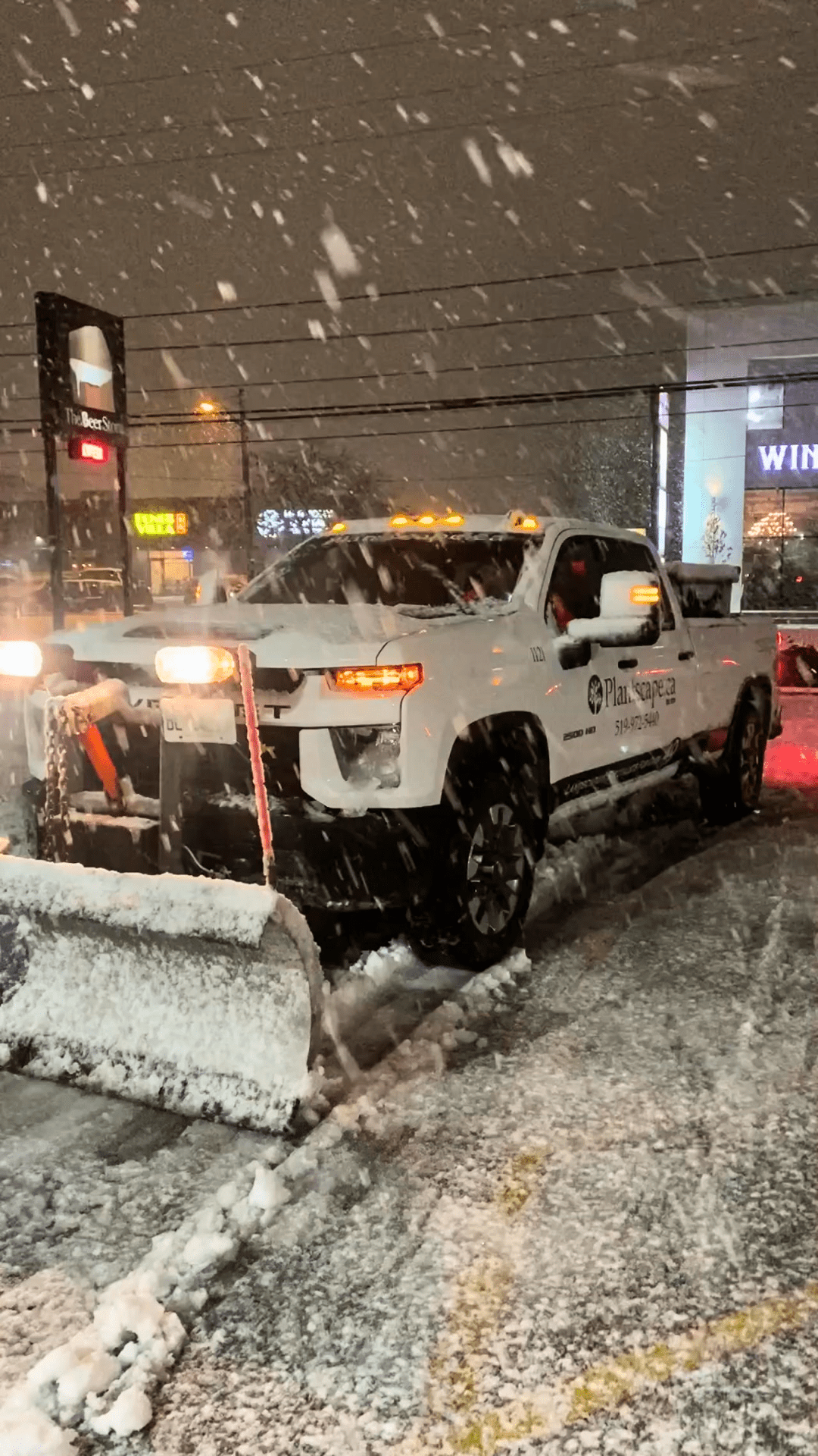 A white pickup truck with company branding is plowing through heavy snow at night, clearing a path near commercial buildings. Snowflakes are visible in the air.