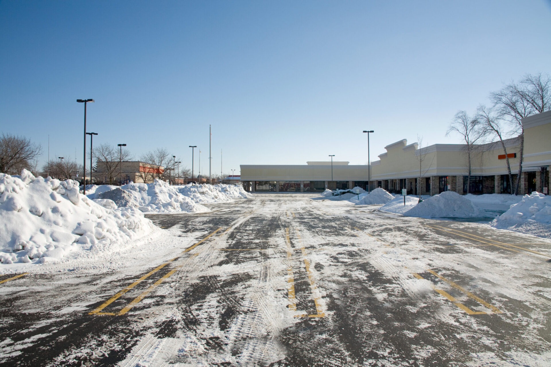 A snowy parking lot with large piles of snow, clear skies, and empty retail storefronts. Tire tracks mark the otherwise white landscape.