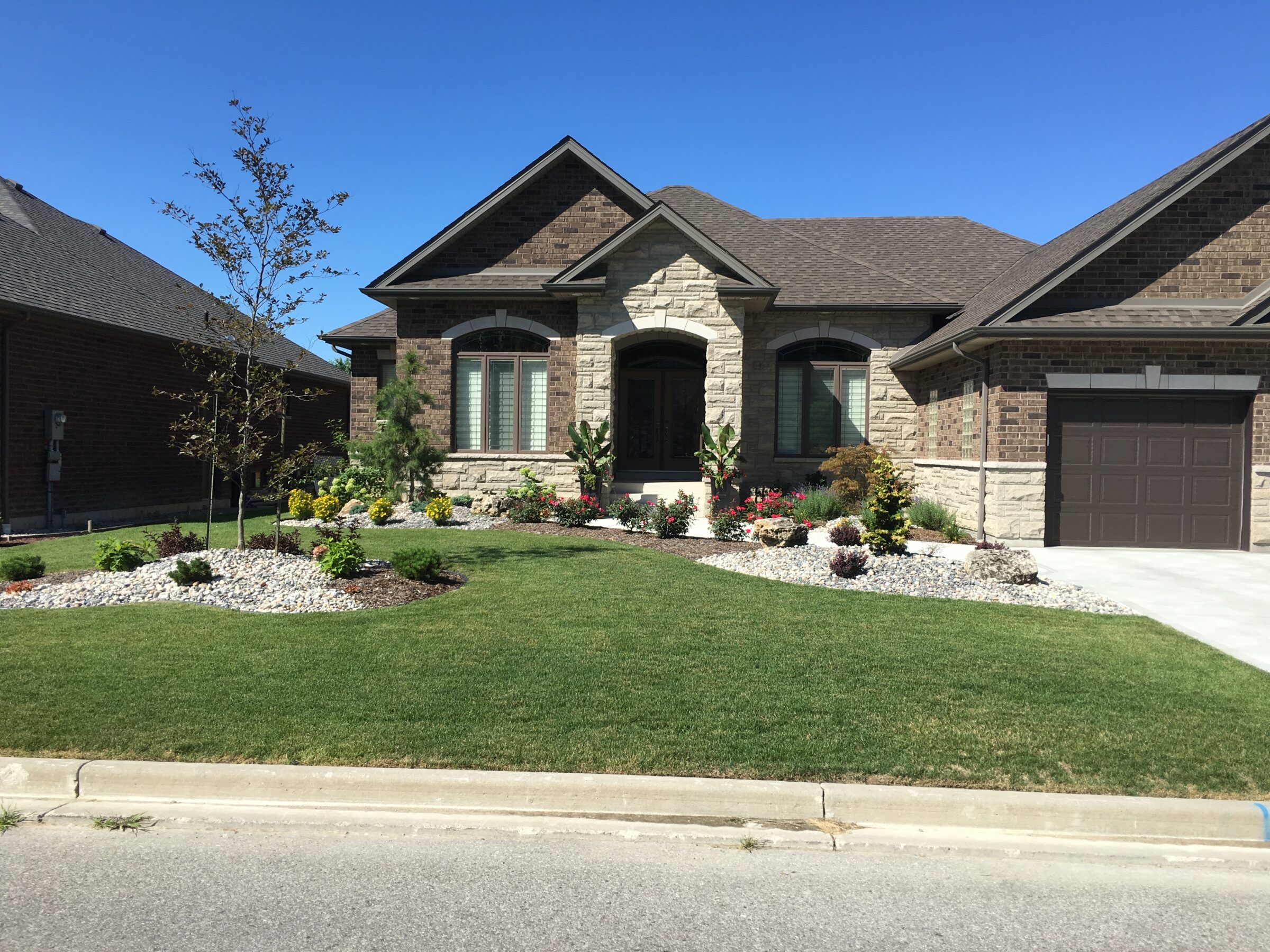 A single-story house with stone facade, well-landscaped garden, green lawn, and a closed single garage door under a clear blue sky.