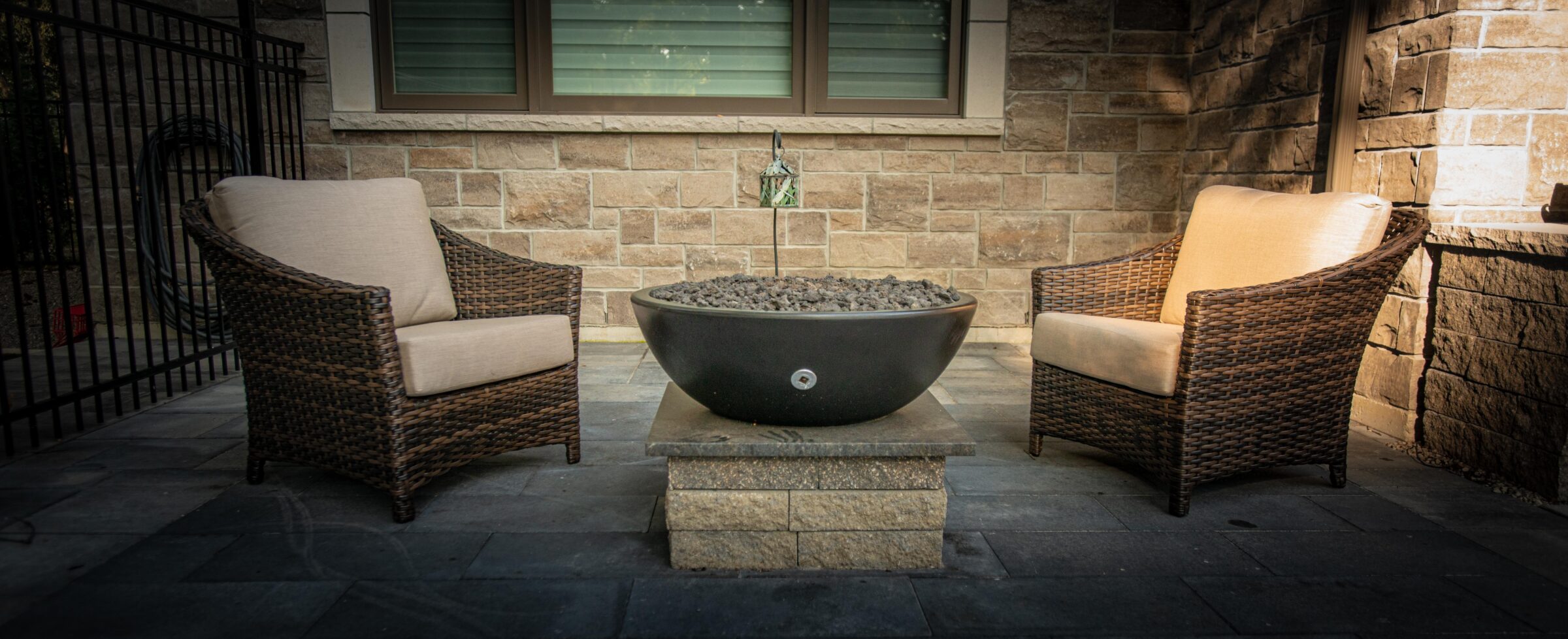 An outdoor patio with two wicker armchairs facing a central fire pit. Neutral cushions, stone tiles, and a brick house backdrop set a relaxing scene.