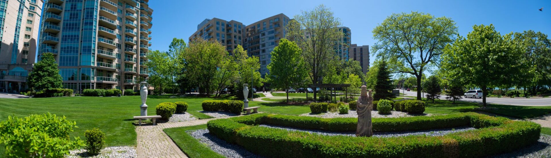 A panoramic view of an urban park with green grass, hedges, trees, sculptures, benches, and surrounding high-rise buildings on a clear day.