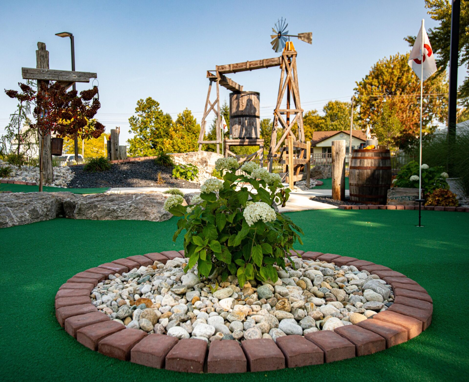 A miniature golf course with green artificial turf, decorative stonework, a wooden windmill, and a white flag on a sunny day.