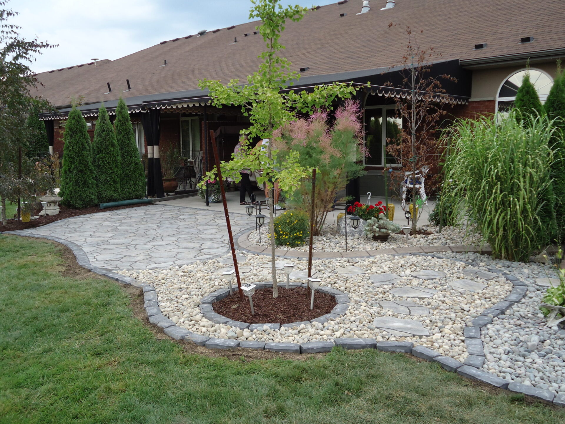 A neatly landscaped backyard features a curved stone patio bordered by white river rocks and mulched garden beds with young trees and ornamental shrubs. The area is adjacent to a brick home with black patio curtains, providing a cozy outdoor living space.
