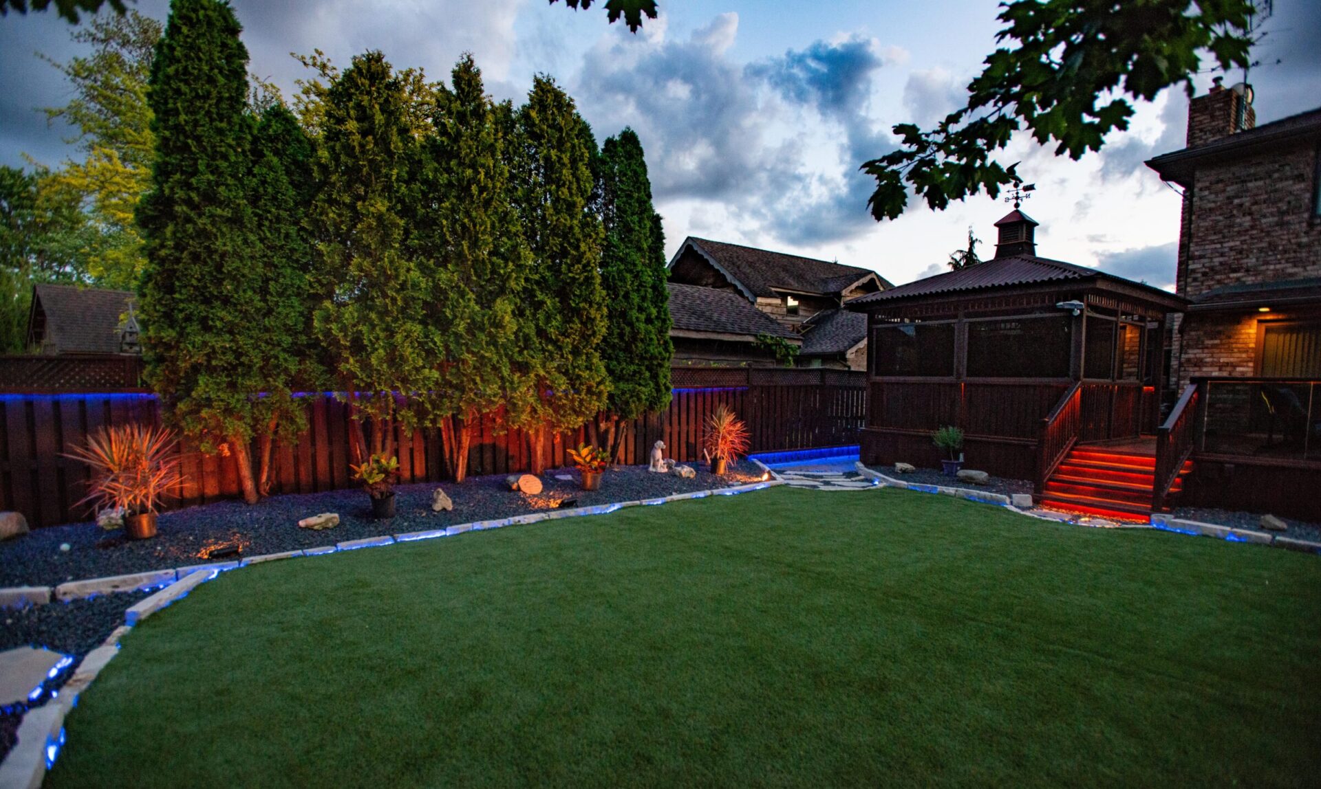 A backyard at dusk features a well-manicured artificial lawn bordered by glowing LED pathway lights and decorative rock beds. Tall evergreens line a dark wooden fence, and red lighting accents the steps of a covered gazebo connected to a brick home, creating a cozy and inviting evening ambiance.