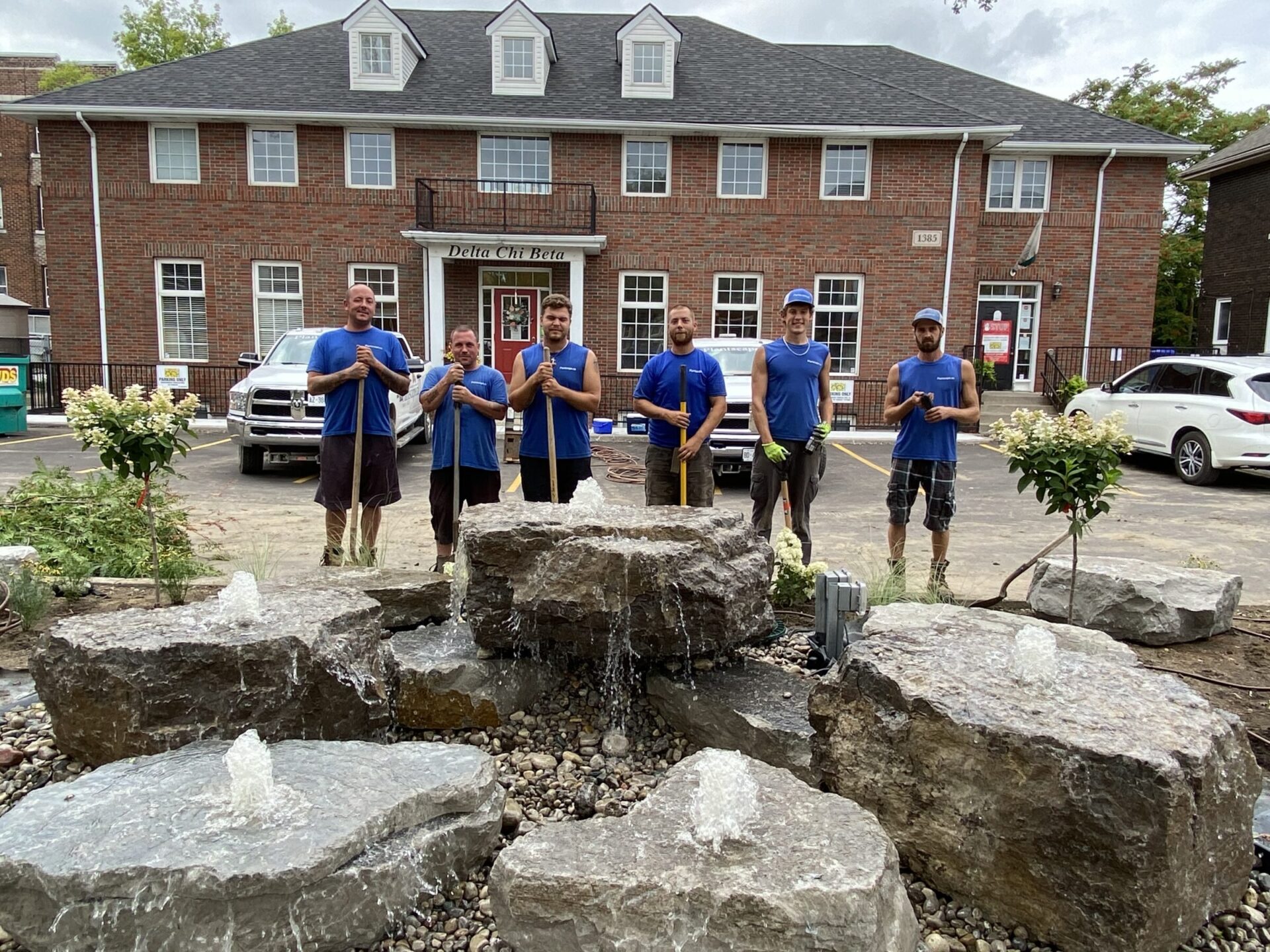 Six men in blue work shirts pose proudly in front of a newly installed rock water feature, with gentle bubbling fountains atop large stone slabs. Behind them stands a red brick building labeled "Delta Chi Beta," and the landscaping around the fountain includes small flowering shrubs and decorative gravel.